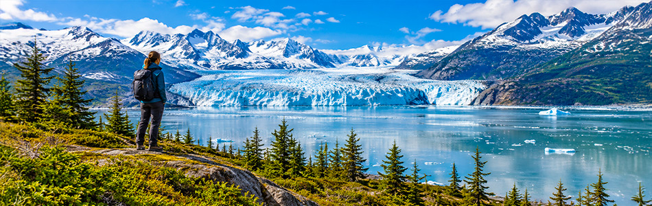 Hiker overlooking a massive tidewater glacier surrounded by Alaska's snowcapped mountains and pristine wilderness.