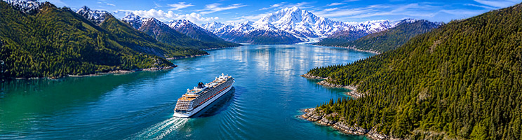 Aerial view of a cruise ship traveling through Alaska with mountain scenery and forested coastline.