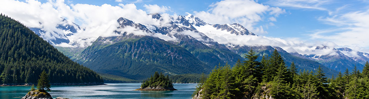 Scenic Alaska fjord surrounded by snow-capped mountains, evergreen forests, and pristine coastal waters.