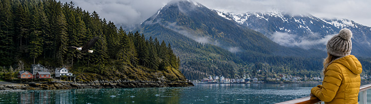 An Alaska cruise traveler watches a bald eagle soar above a forested coastal town surrounded by misty mountains. An Alaska cruise traveler watches a bald eagle soar above a forested coastal town surrounded by misty mountains.